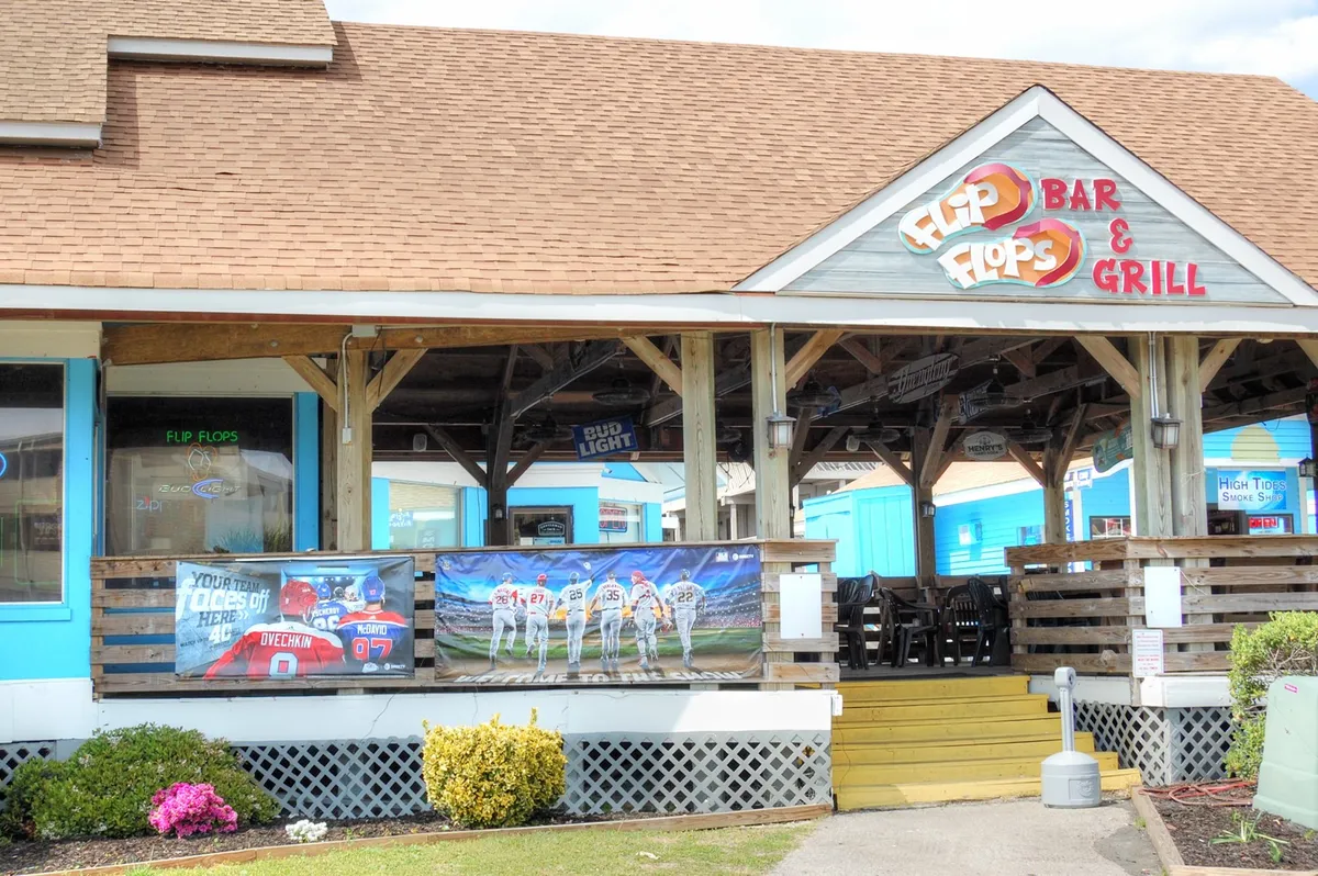 The colorful sign and outdoor seating of Flip Flops Bar & Grill, bordered by sports-themed banners and a cheerful patio area. It’s a welcoming spot for outdoor dining and socializing, tying into the lively local scene.
