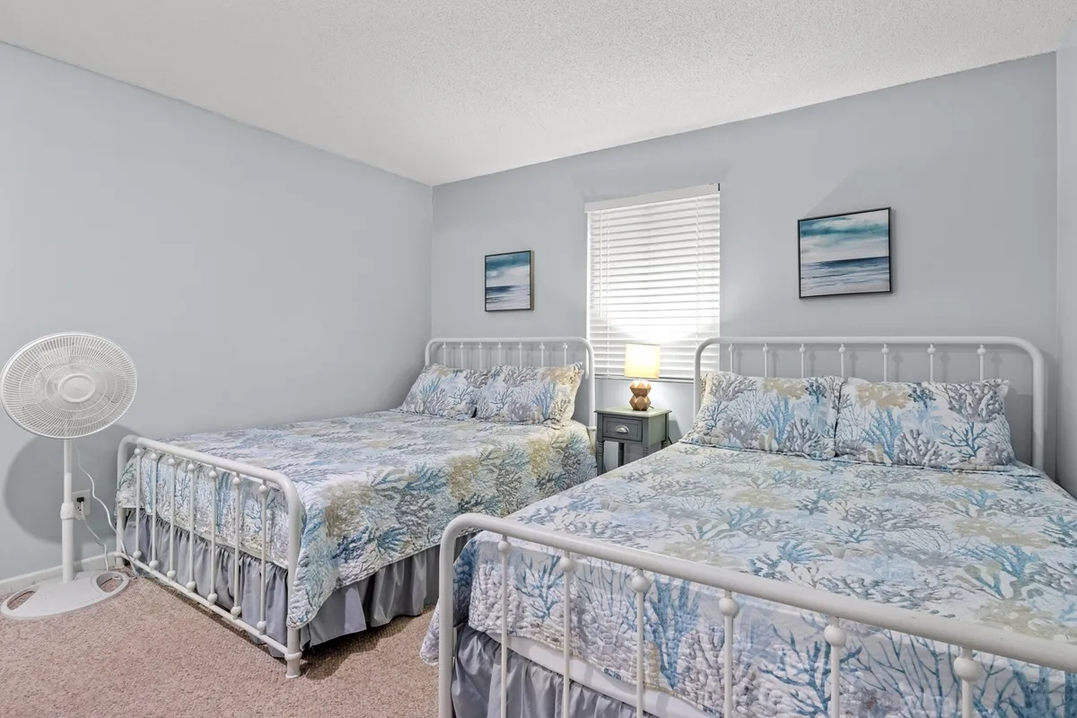 A bright guest bedroom with two queen beds featuring floral bedding, a nightstand with a lamp for cozy reading sessions, and elegant wall art. The arrangement offers both comfort and functionality—ideal for families, friends, or golf buddies traveling together.