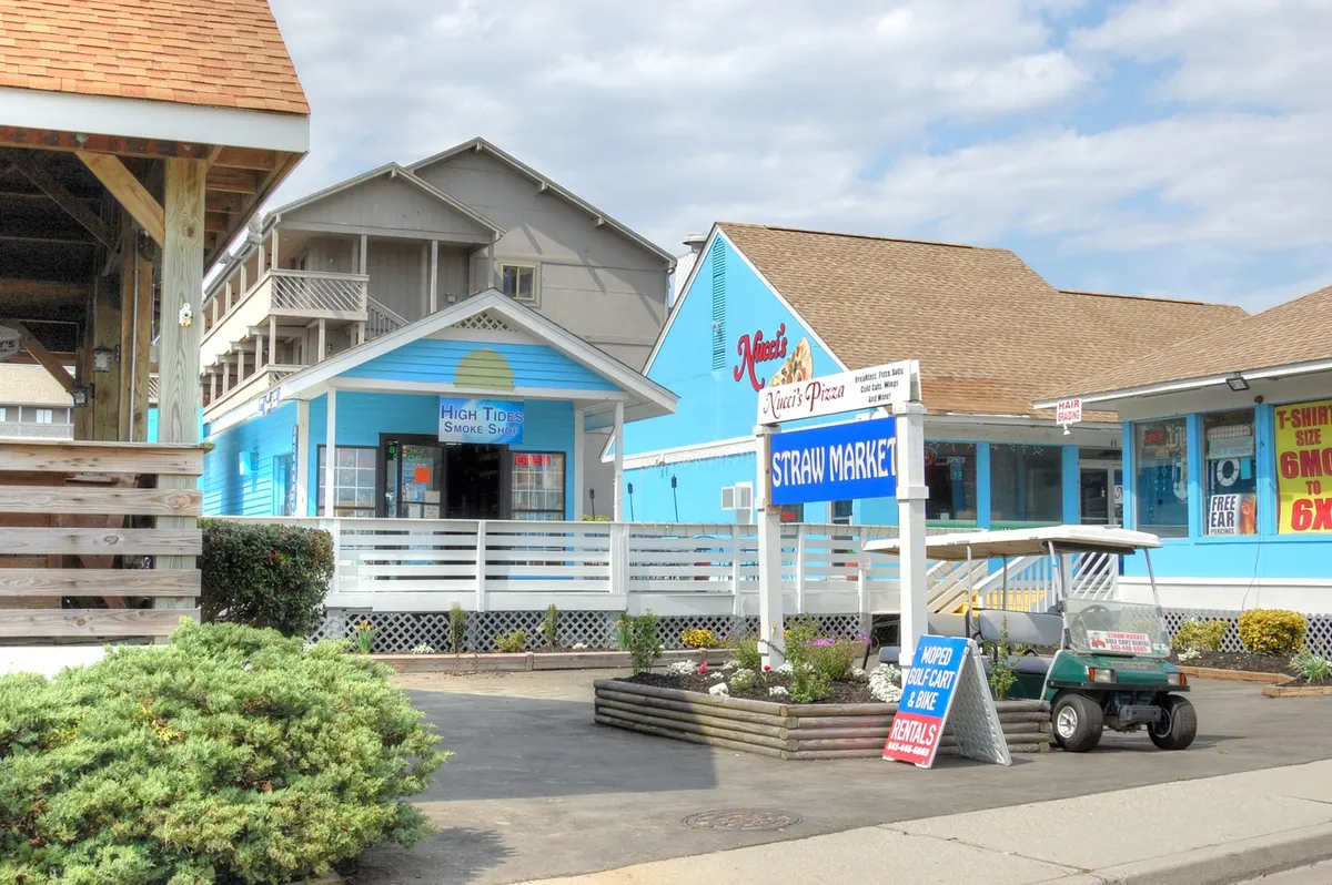Colorful storefronts line a sunlit walkway, with a golf cart parked nearby and prominent signage for Straw Market and Meek's Pizza. The cheerful mood suggests a day spent shopping, dining, or enjoying a sunny outing close to home.