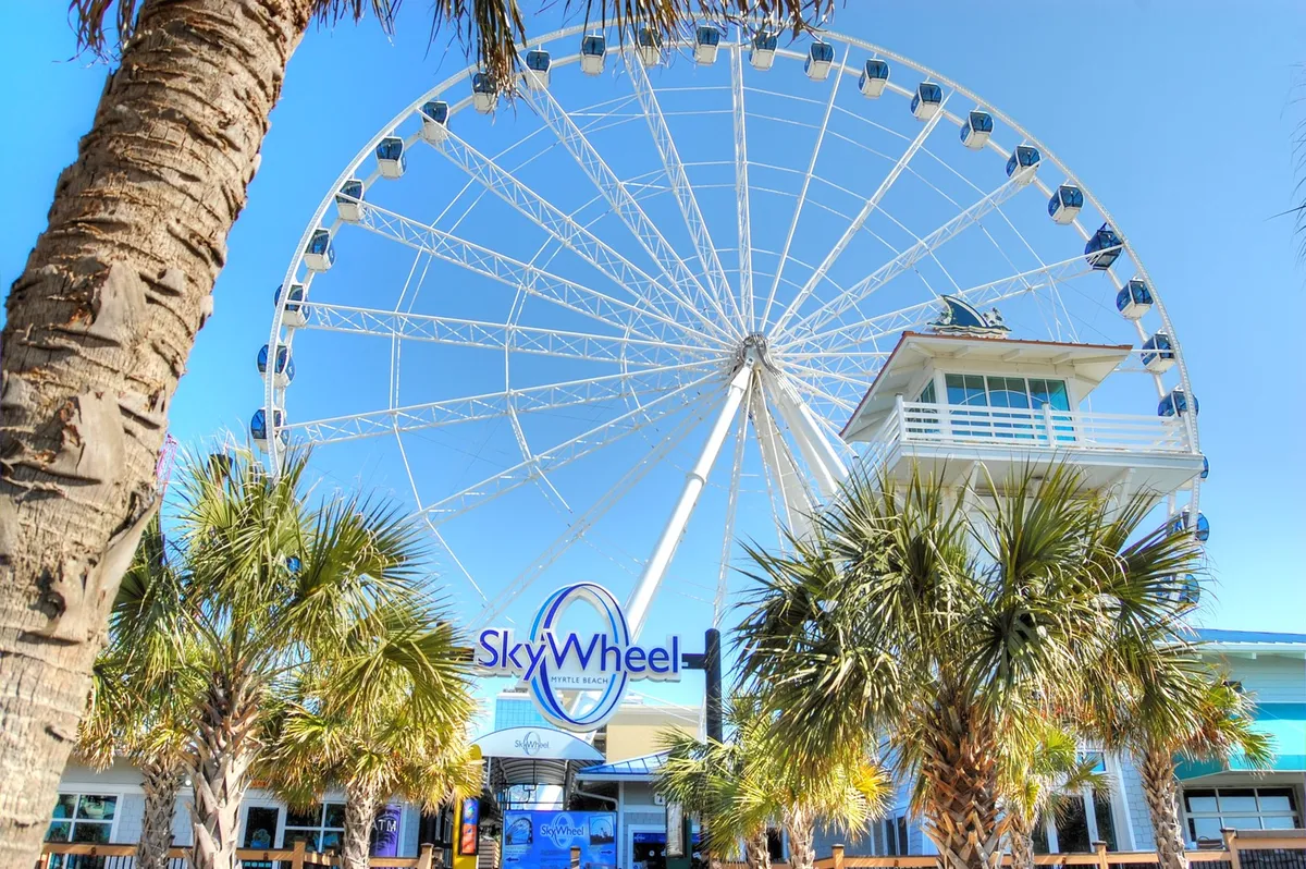 This image features the iconic Myrtle Beach SkyWheel, palm trees, and the entrance building, all under a bright sky. The scene is lively, highlighting one of the region's top attractions included in the Free Fun Program. The setting conveys excitement and the promise of memorable experiences.