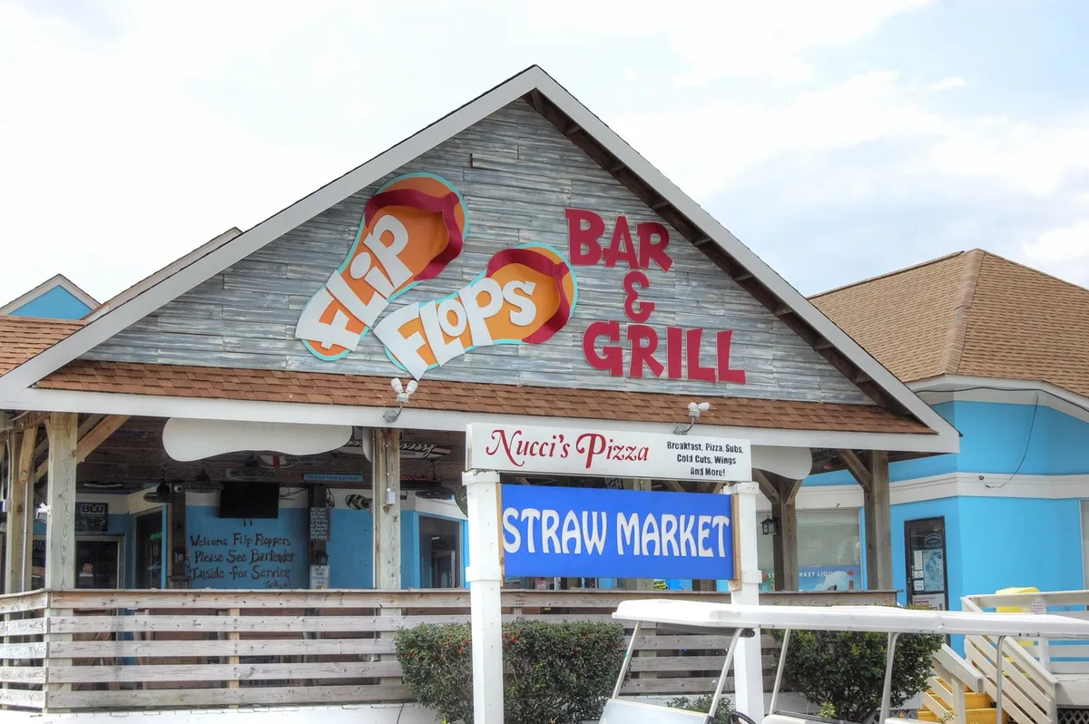 A lively exterior scene featuring the Flip Flops Bar & Grill, Nucci's Pizza, and Straw Market, with signage indicating options for dining, drinks, and local exploration. The casual setting suggests a vibrant local nightlife and post-golf hangout spots.