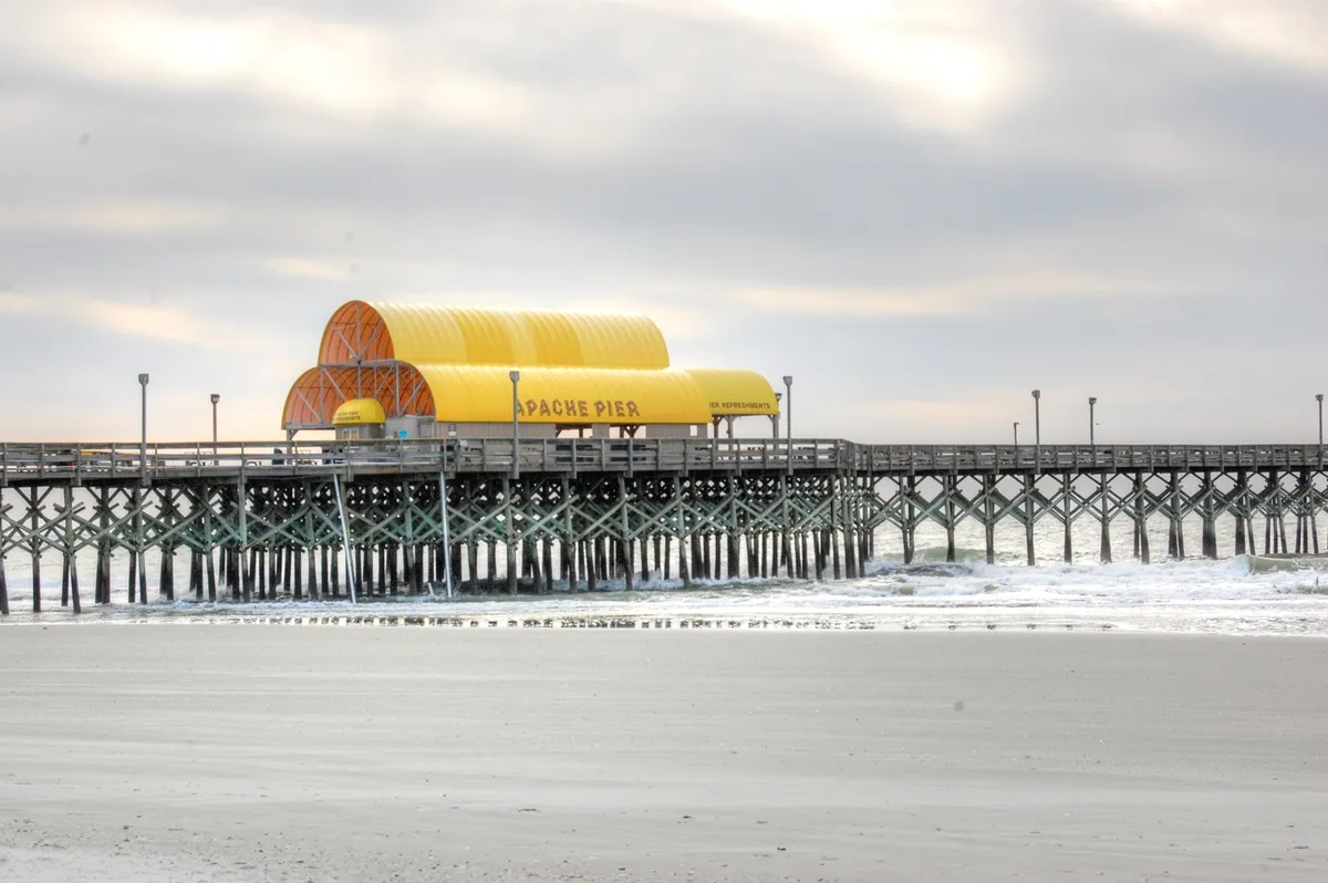 A sunlit view of Apache Pier with its signature yellow roof, sturdy supports stretching out over the gentle waves, and a sandy beach in the foreground. The relaxed oceanfront vibe is enhanced by people visible along the pier, enjoying fishing and family outings.