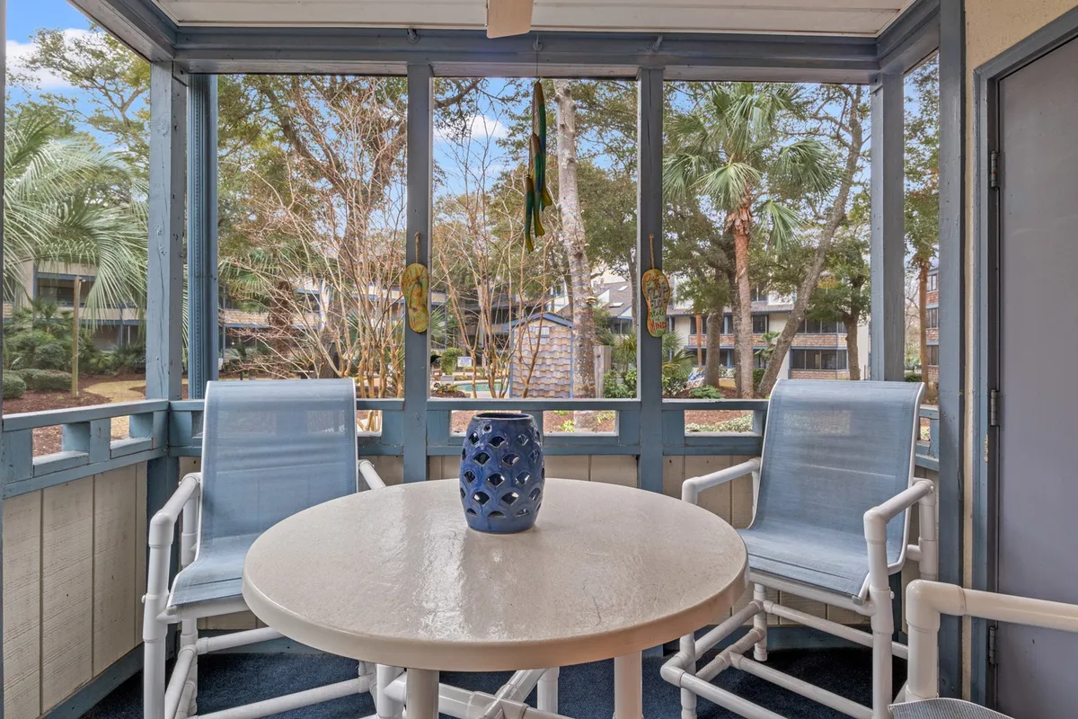 A tranquil screened-in porch with a table featuring a blue vase, surrounded by lush greenery. The porch is airy and spacious, making it an inviting spot for relaxing, socializing, or storing beach gear after an active day outdoors.