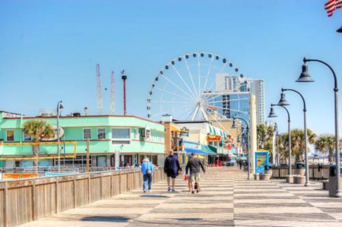The scene shows a bustling Myrtle Beach boardwalk, colorful beachfront buildings, a ferris wheel, and people enjoying a stroll. The atmosphere is joyful and inviting, suggesting a hub of entertainment options and family-friendly fun, perfectly aligning with the narrative of daily complimentary activities.