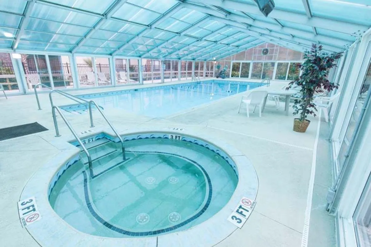 This image shows an inviting indoor swimming pool with a hot tub and ample seating around the pool area. Large windows let in natural light, enhancing the tranquil ambiance and showcasing the space's openness and connection to the outdoors.