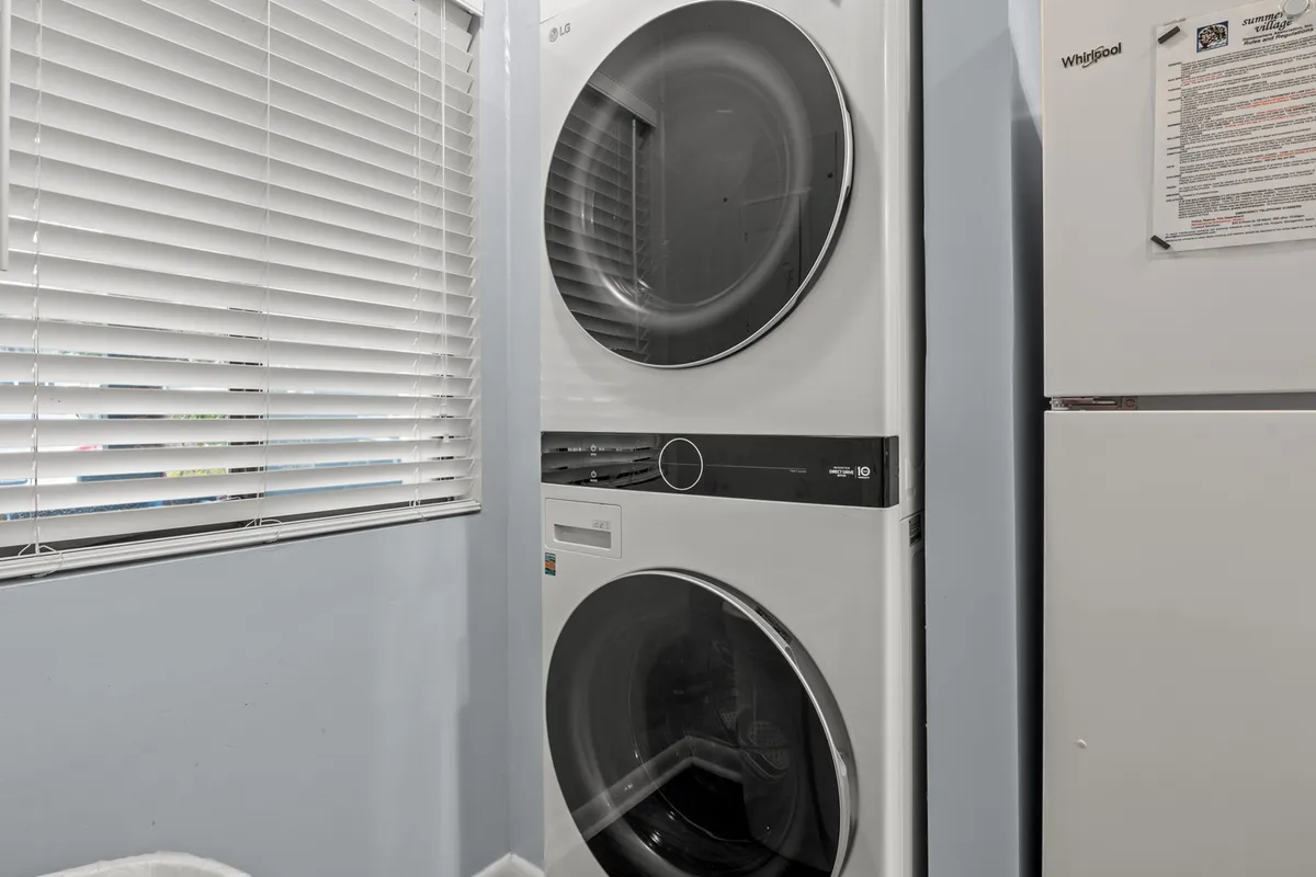 A tidy laundry area featuring a stacked washer and dryer next to a white refrigerator, with blinds filtering soft natural light. The space is efficient and functional, supporting the needs of an active vacation.