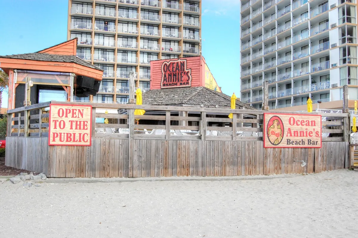 The lively exterior of Ocean Annie's Beach Bar, marked by its welcoming sign, yellow umbrellas scattered across the deck, and beachgoers relaxing nearby. The ocean is just steps away, adding to the casual, sun-kissed atmosphere.